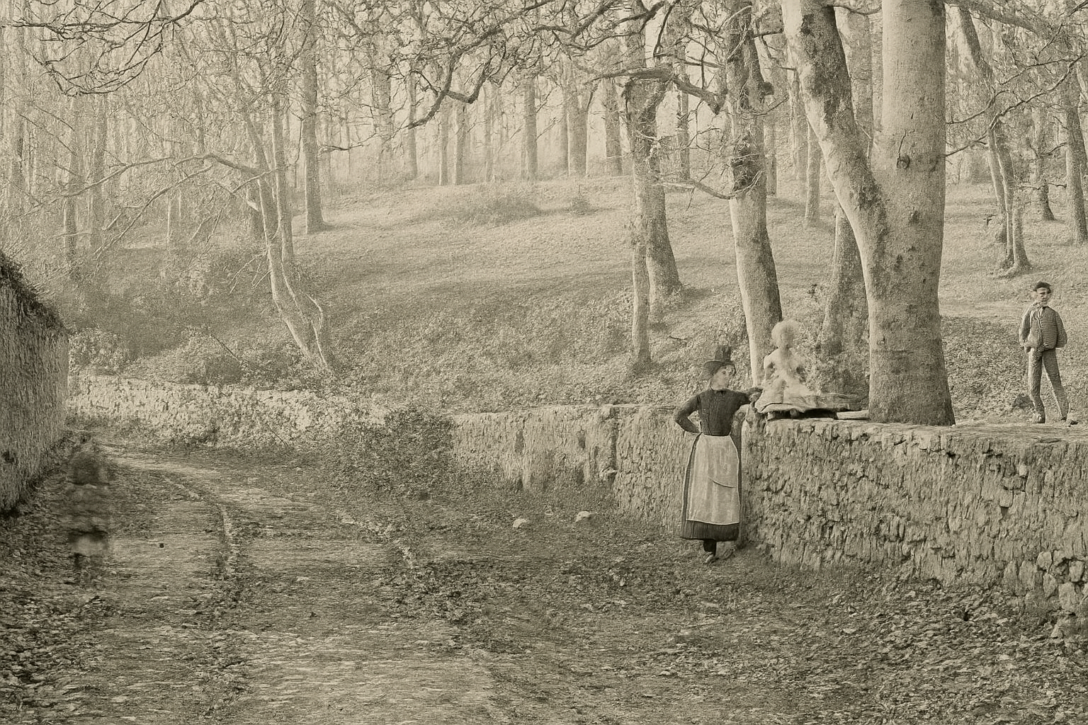 Photograph c.1860: woodland slope and dry stone wall at Rushen Abbey, with a young woman in working apron, a small child on the wall, and a boy in the distance.